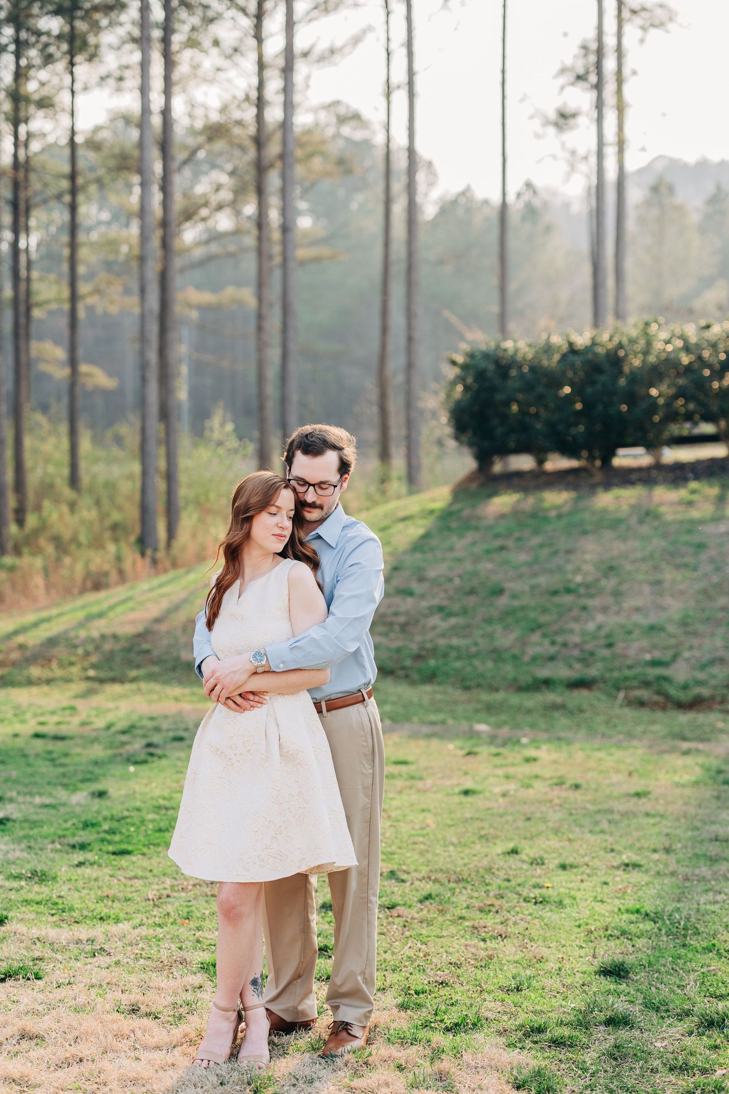 A couple embraces in a romantic outdoor setting with tall pine trees and grassy hills in the background at Clisby Pines