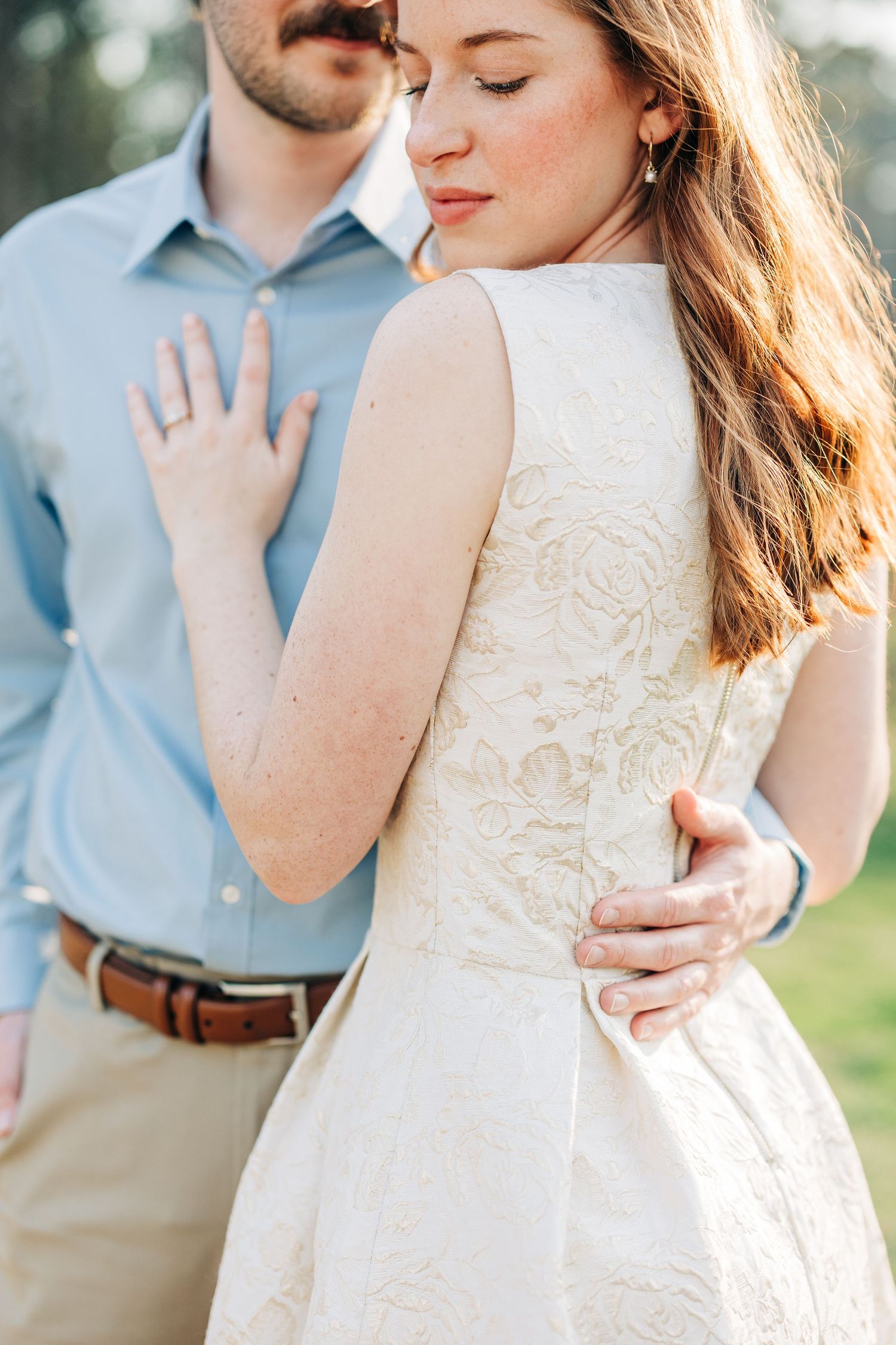 A romantic couple embraces in soft sunlight wearing a white lace dress and light blue shirt during an outdoor photoshoot.
