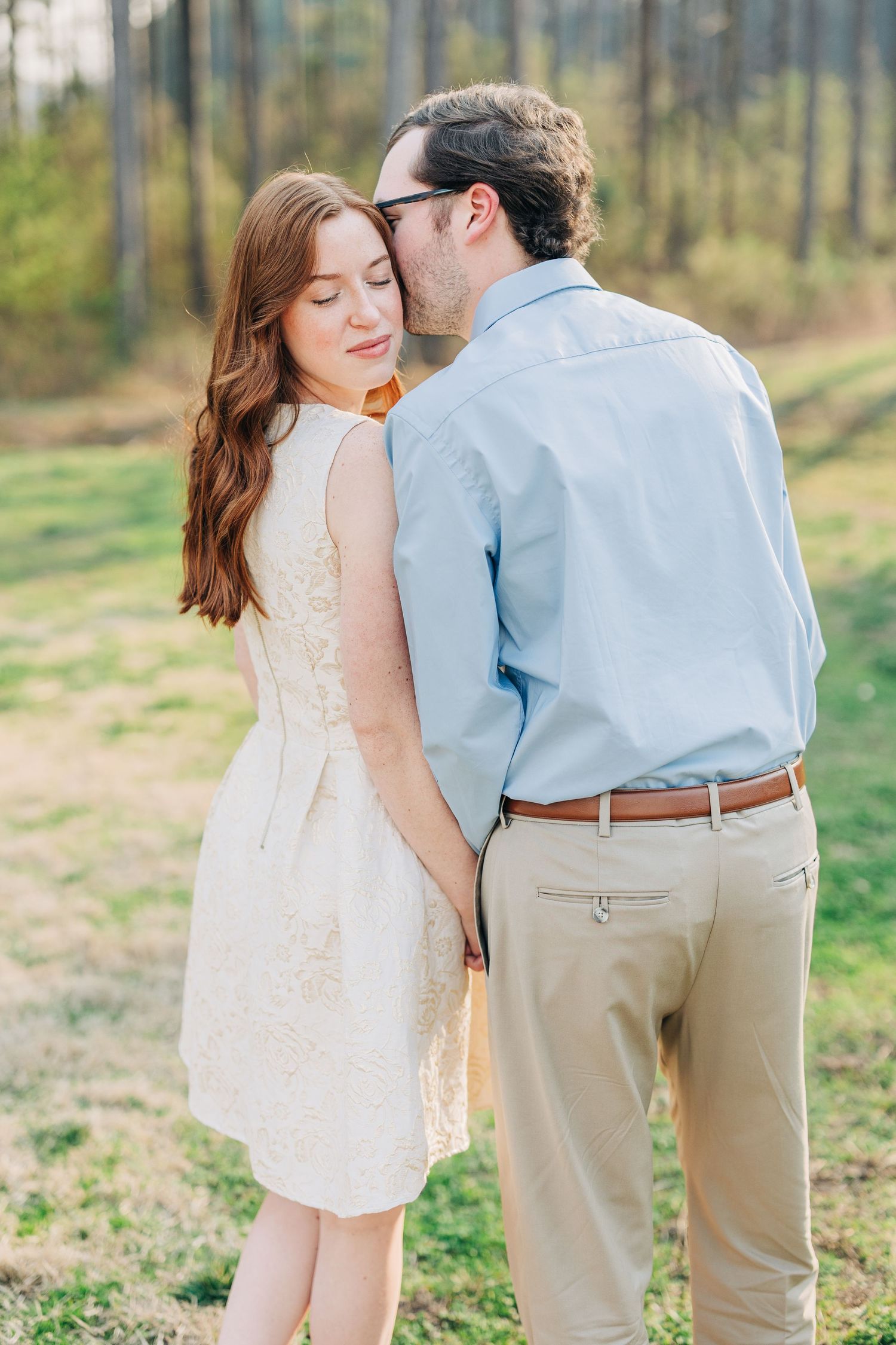 A romantic couple in casual spring attire embraces during an outdoor photoshoot in a sunlit wooded area.