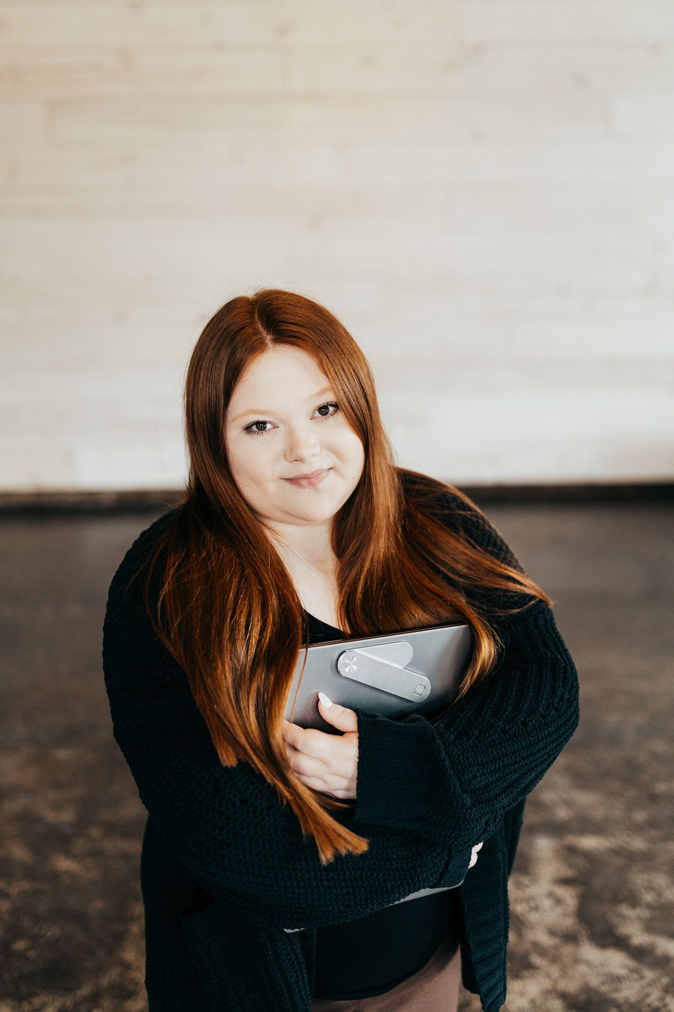 Person with long red hair wearing black sweater holds laptop in creative indoor photo series.