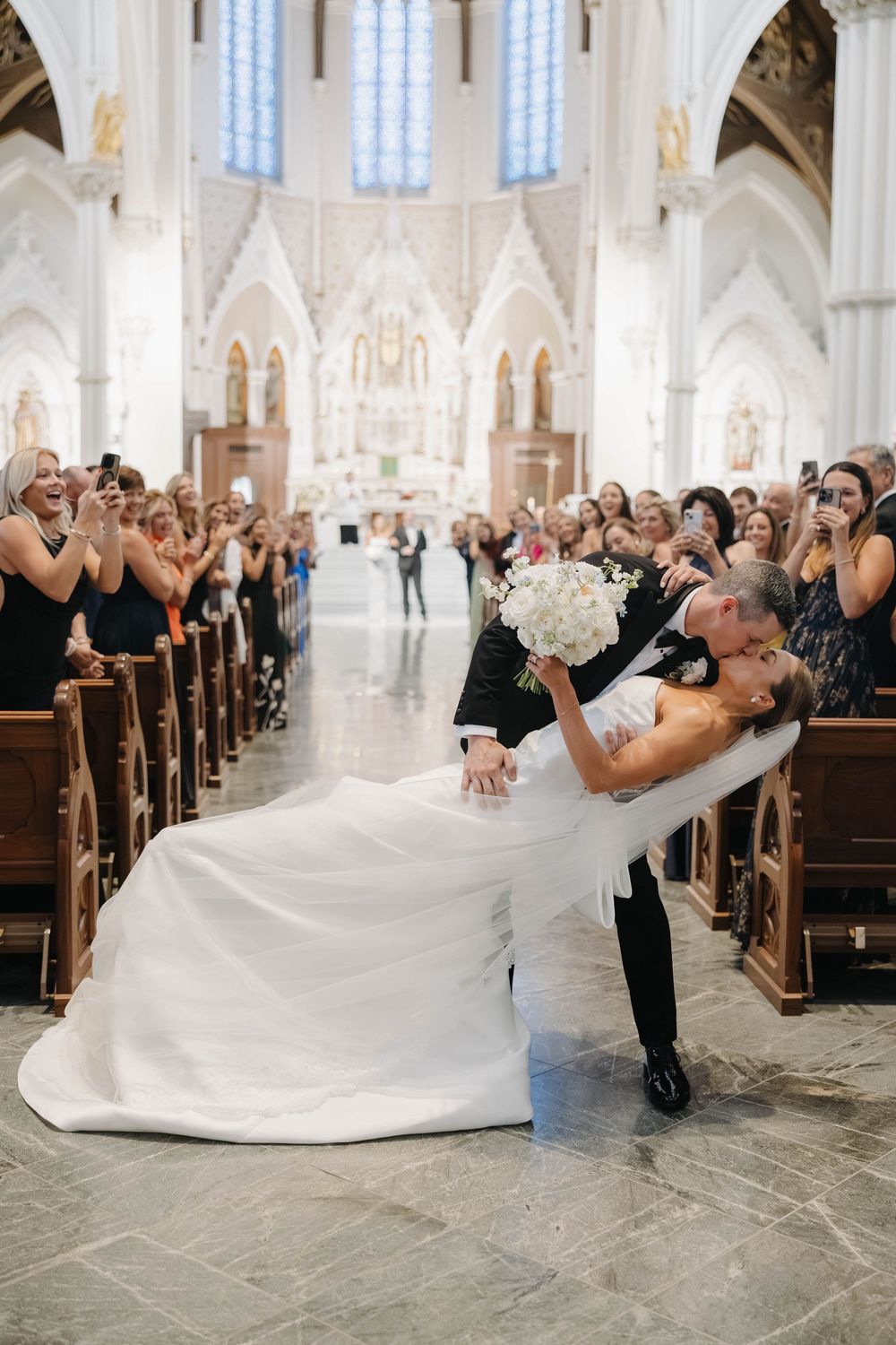 A romantic dip during a wedding ceremony in a grand cathedral with white decor and high arched ceilings.