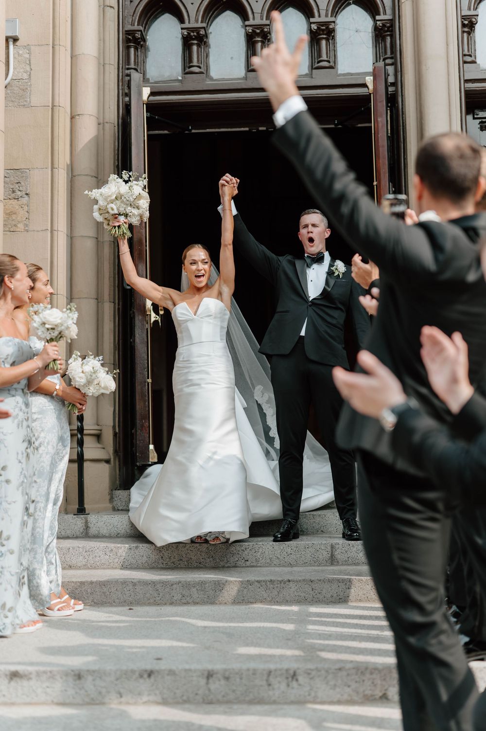 Wedding party celebrates joyfully outside a grand stone church entrance with raised arms and white bouquets.