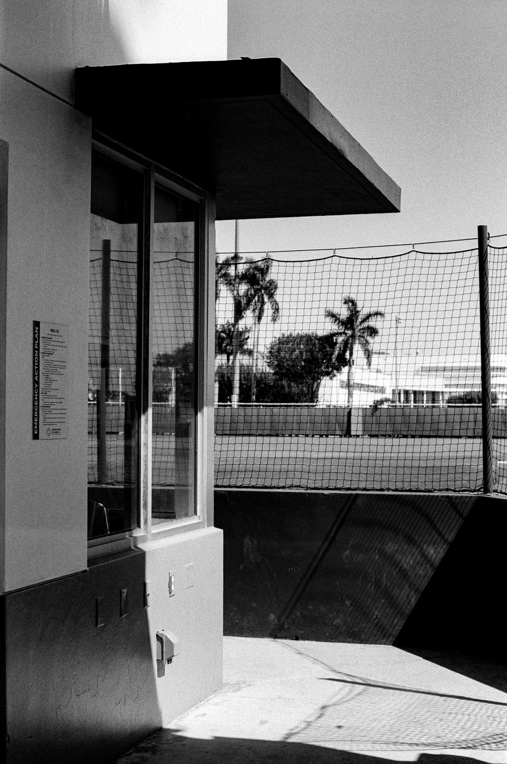 Black and white architectural photo showing a building entrance with overhang, chain link fence and palm trees in background.
