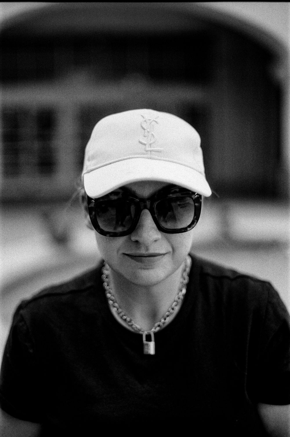 Black and white portrait of someone wearing sunglasses, a baseball cap, and a black t-shirt with a chain necklace.