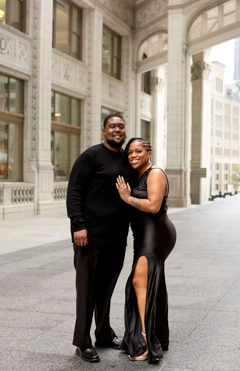 Elegant couple in matching black outfits posing in front of a grand architectural building in the city.
