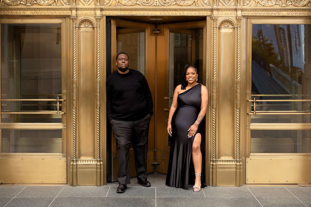 Elegant couple in matching black outfits posing before ornate gold Art Deco doors.
