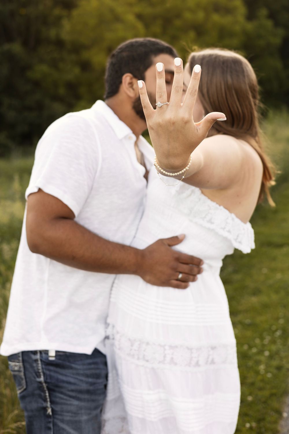Engaged couple kissing outdoors while woman displays diamond engagement ring toward camera.