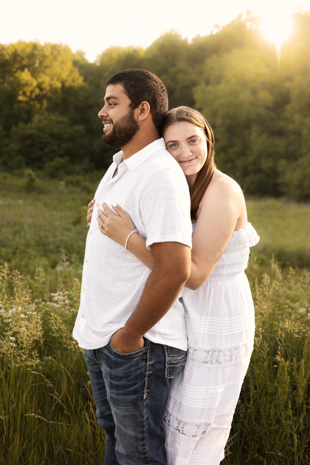 Happy couple embracing in a sunlit field, woman in white dress hugging man from behind at golden hour.