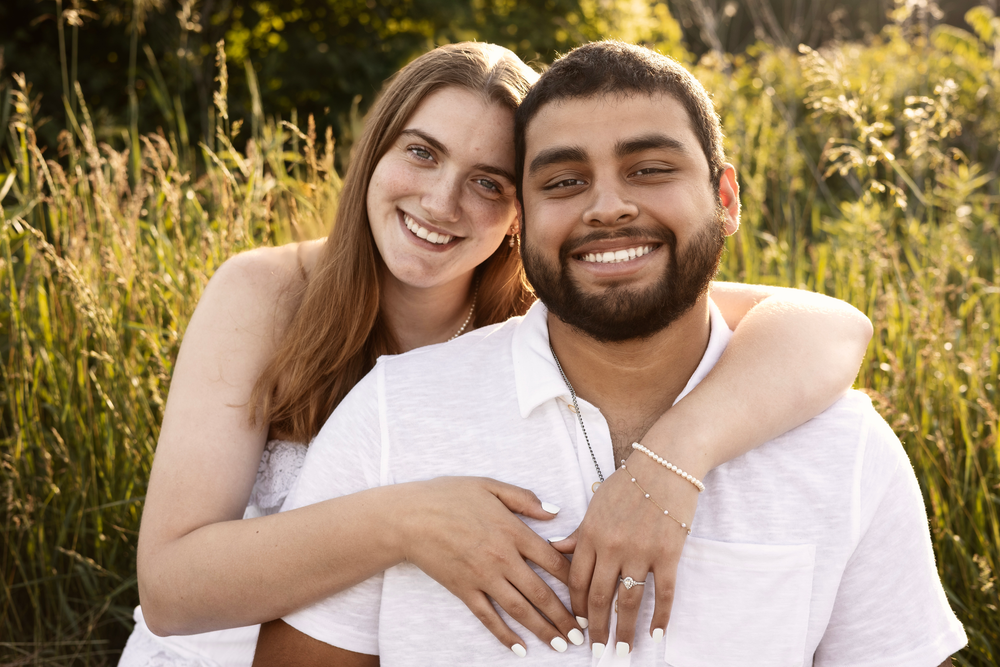 Joyful engaged couple sitting outdoors in matching white outfits, displaying engagement ring while smiling.