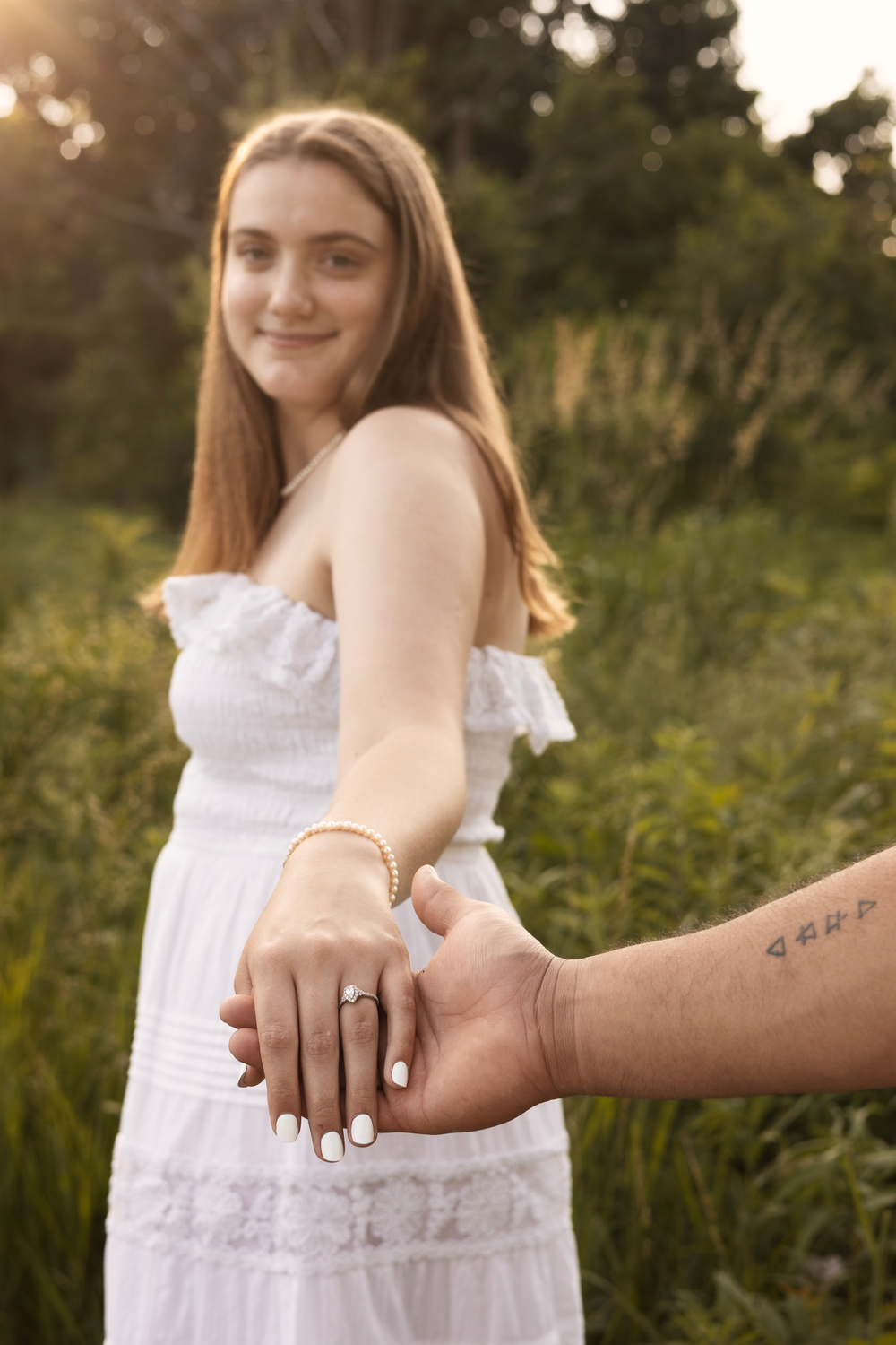 Bride-to-be in white dress showing off her diamond engagement ring while holding her partner's hand outdoors.
