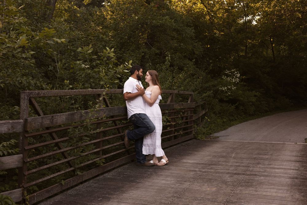 Romantic couple sharing an intimate moment on a wooden bridge surrounded by lush green trees at dusk.