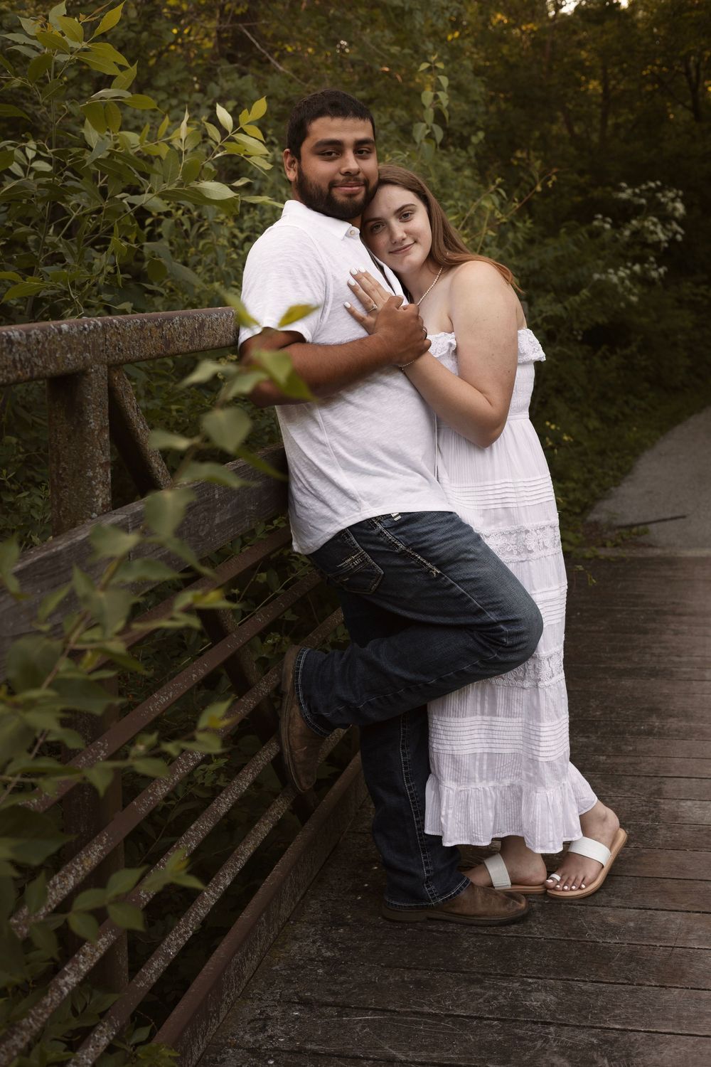 Couple posing on a wooden bridge in white outfits, leaning against the railing surrounded by greenery.