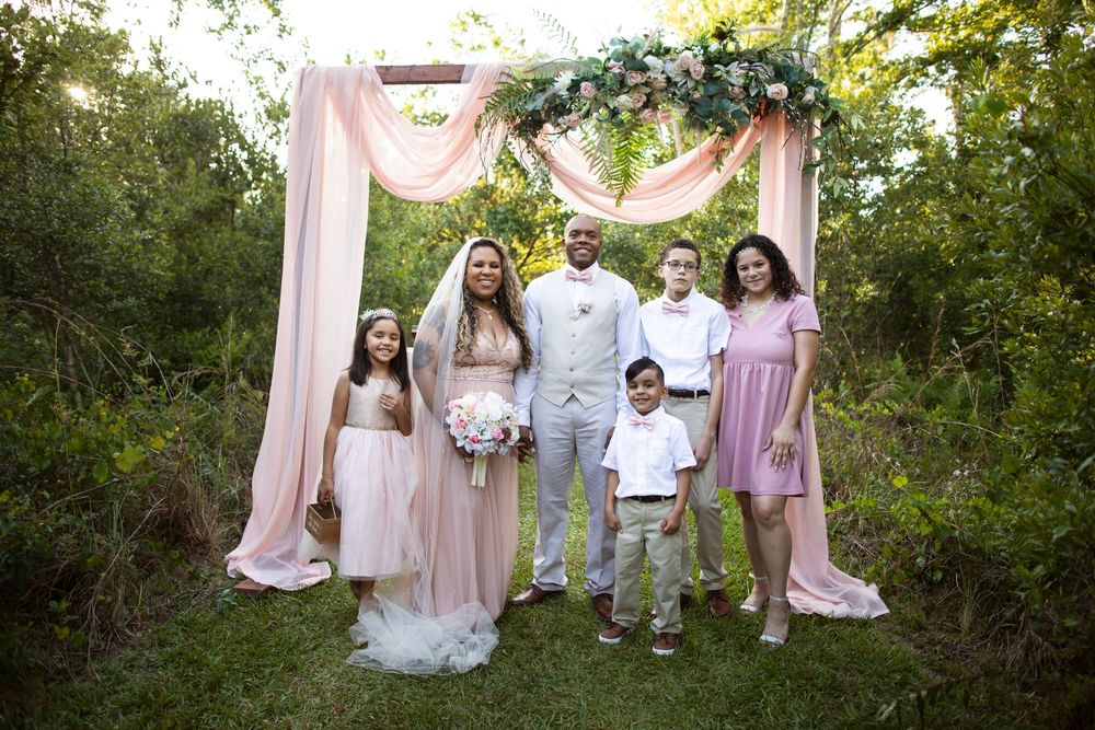 Wedding party in light pink and white attire poses together in a lush garden setting during outdoor celebration.