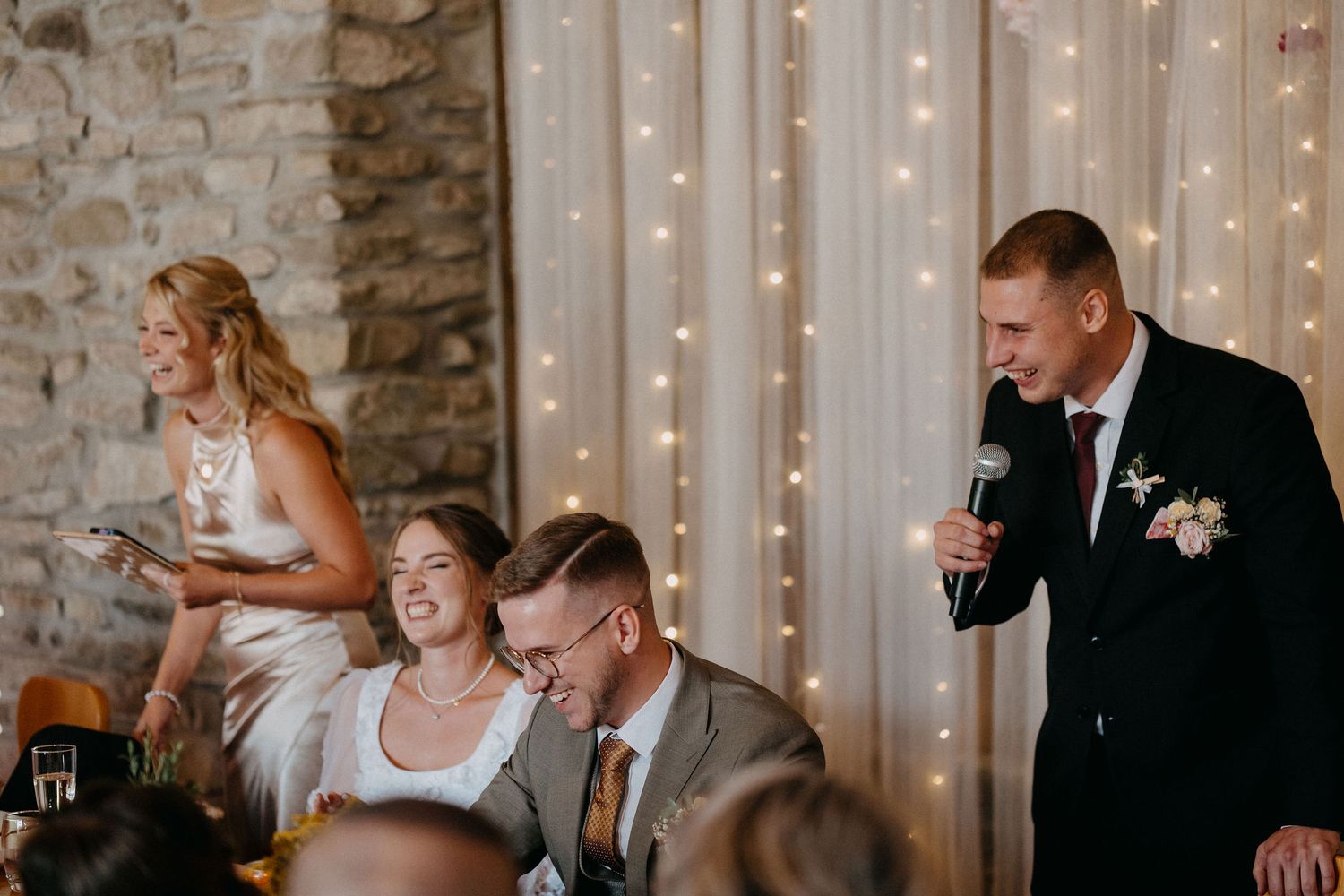 Wedding reception guests enjoying a candid moment at a table with twinkle lights and stone wall backdrop.