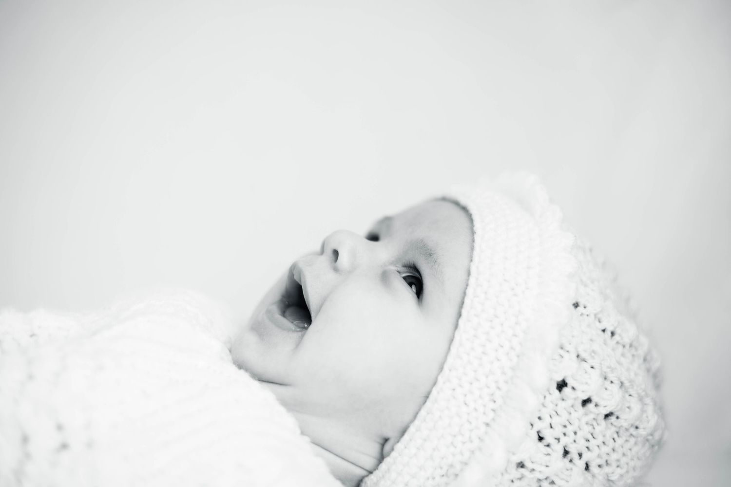 Black and white portrait of baby in white knit bonnet looking up with mouth open.