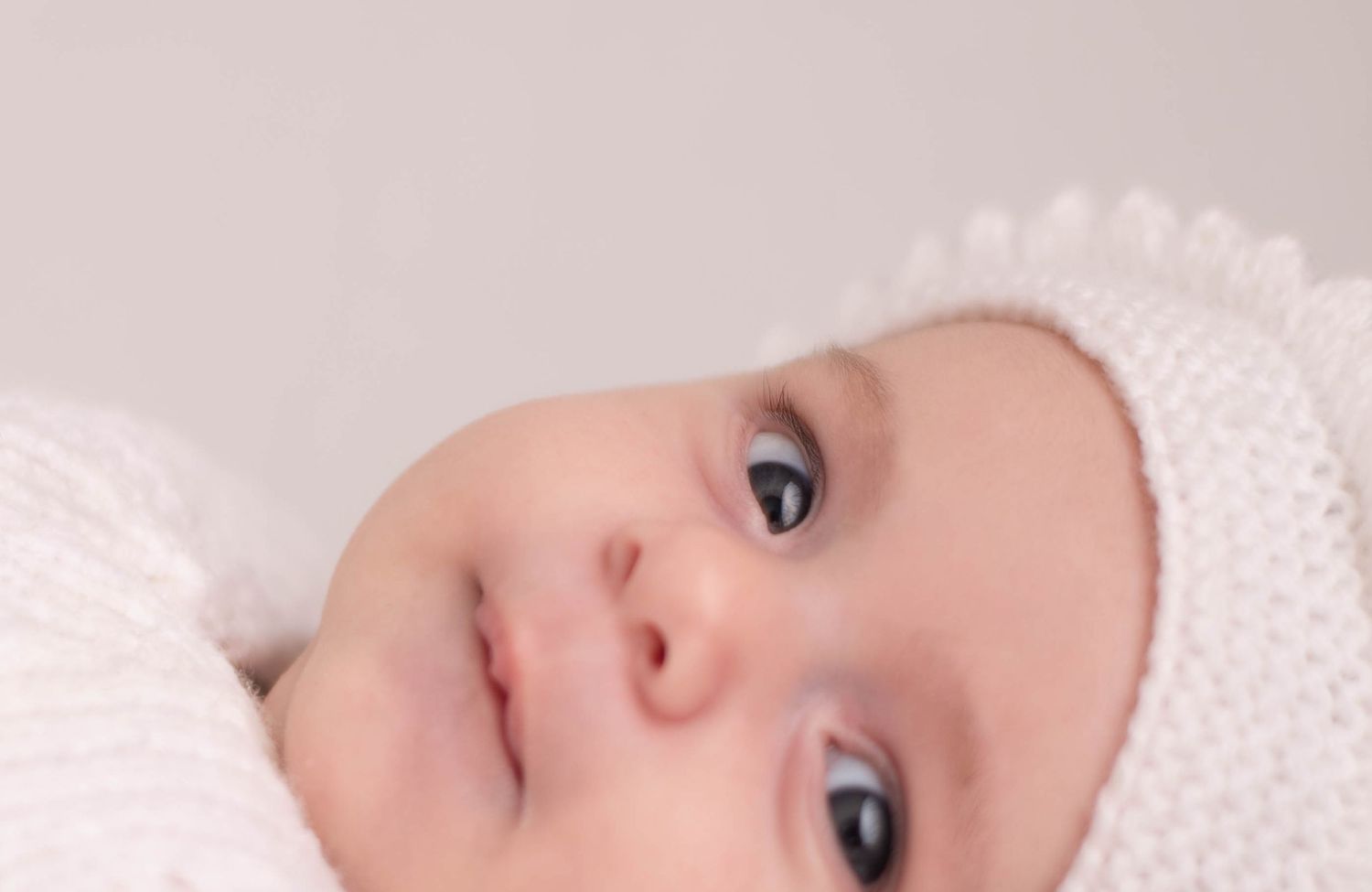 Close-up portrait of baby with blue eyes wearing white knit bonnet and soft wrap.