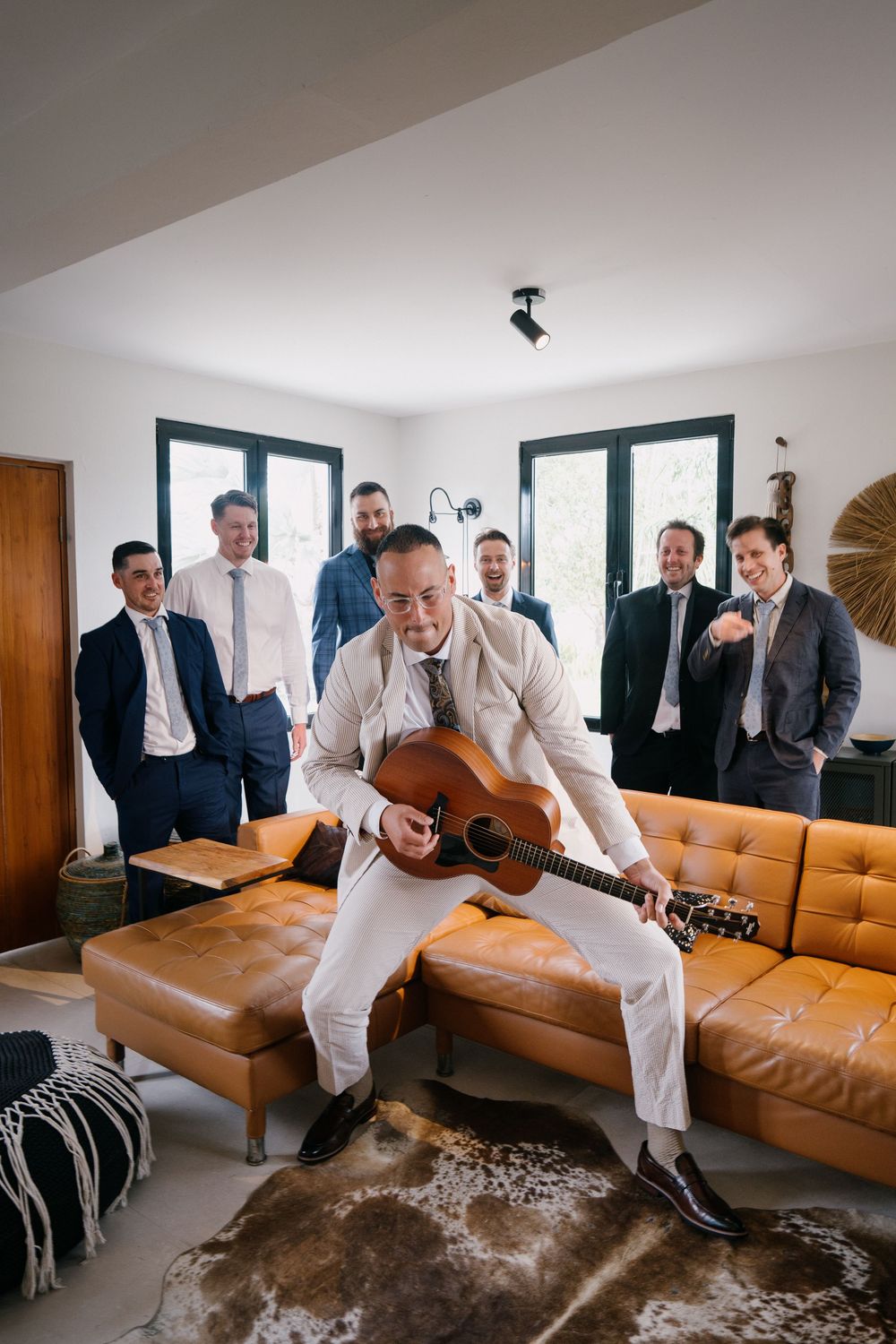 A sequence of photos showing men in white suits jumping on a brown leather couch in a bright modern room.