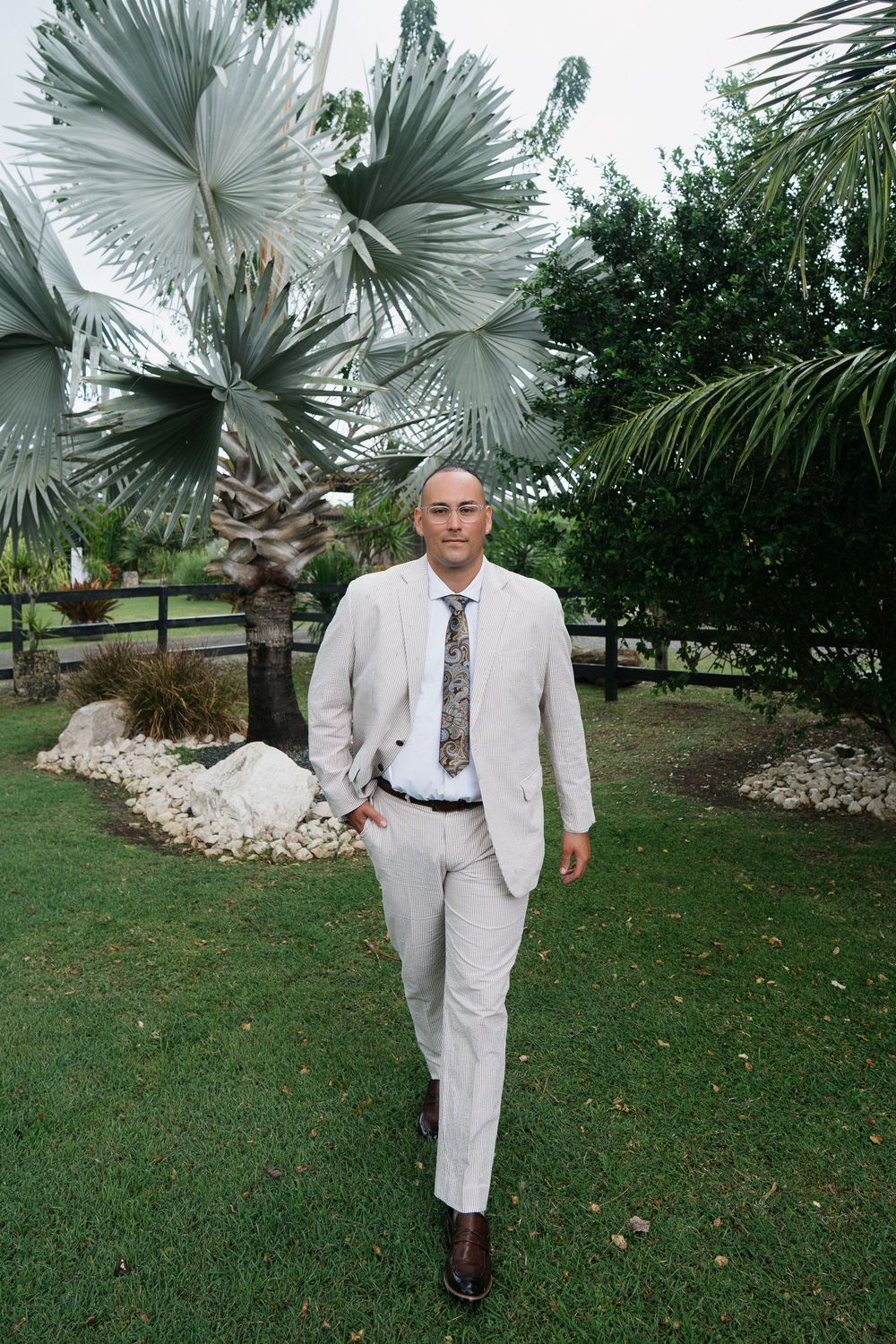 Someone in a light-colored suit walks on green grass with palm trees and landscaping in the background.