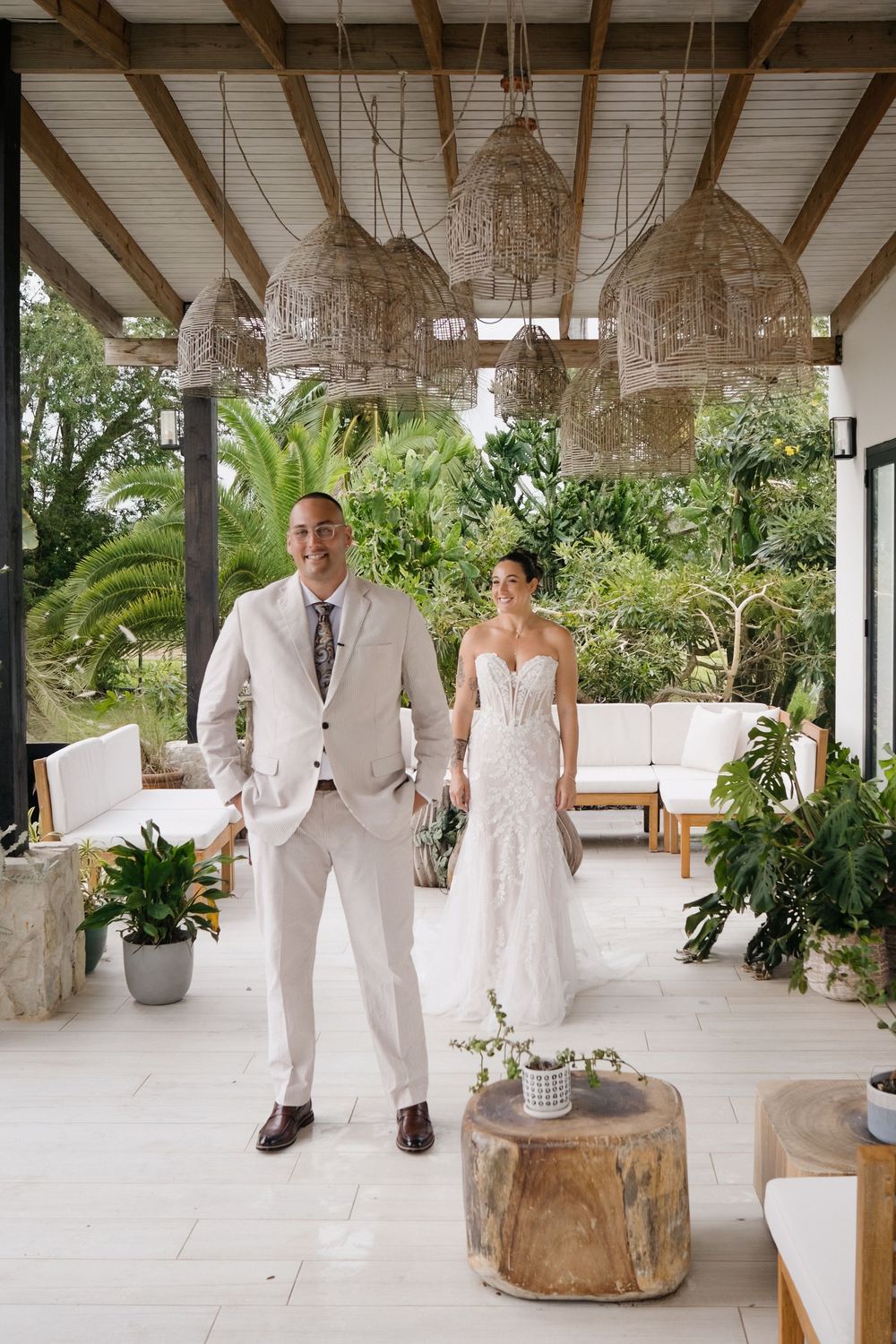 Wedding couple in white attire poses in an elegant outdoor venue with crystal chandeliers and tropical greenery.