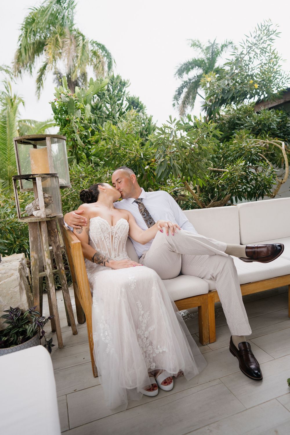 Couple in white wedding attire relaxing on a wooden bench surrounded by tropical plants on a patio.