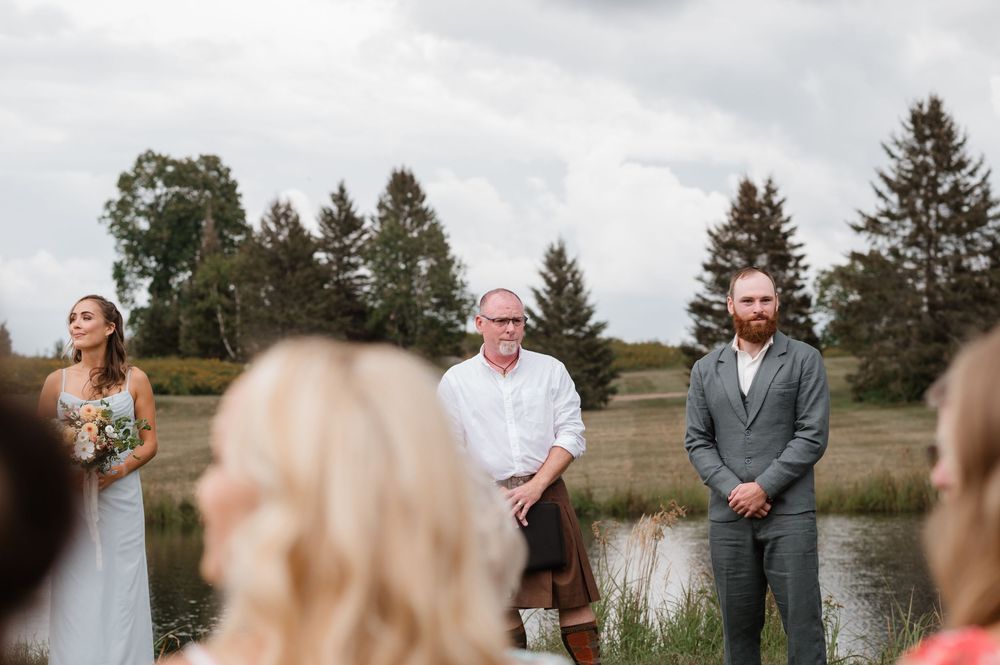 Wedding party stands in a grassy field on an overcast day while guests look on in the background.