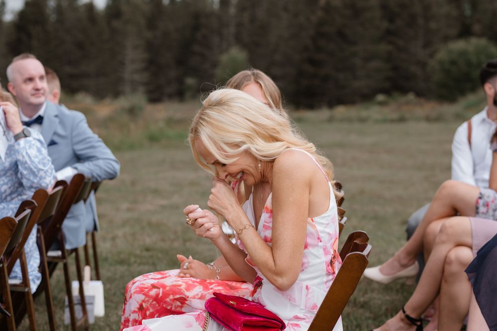 Wedding guests enjoy outdoor reception in rustic wooden chairs arranged in rows on grassy field.