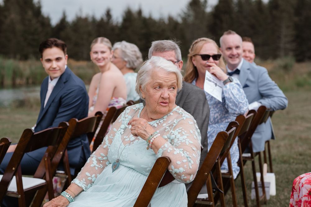 Wedding guests seated in wooden chairs outdoors smile and laugh during an outdoor ceremony in a forest setting.