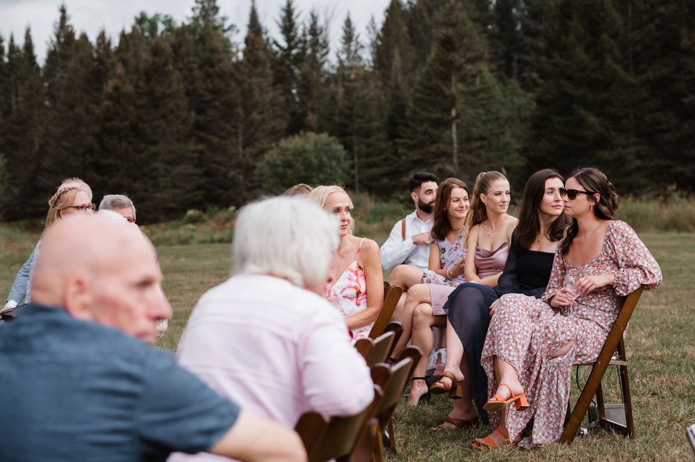 A group of people sitting in chairs on a grassy field surrounded by evergreen trees during an outdoor gathering.
