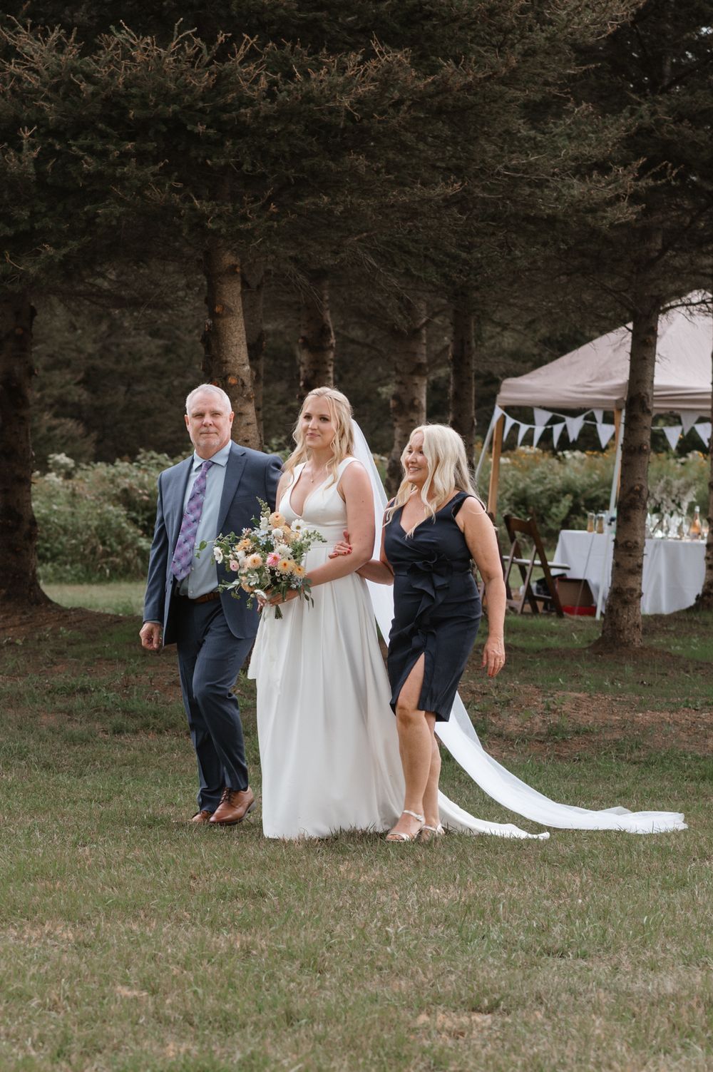 Wedding party members gather outdoors at a rustic ceremony venue with pine trees and decorative banners in the background.