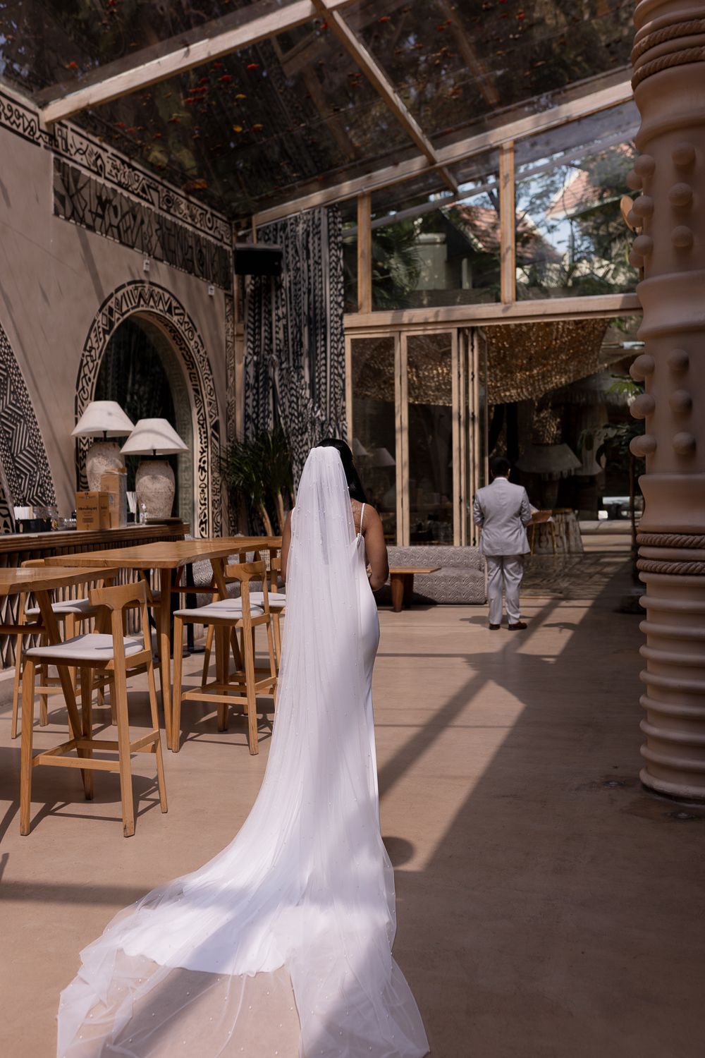 A flowing white wedding dress on display in a covered outdoor patio area with wooden furniture and ornate columns.
