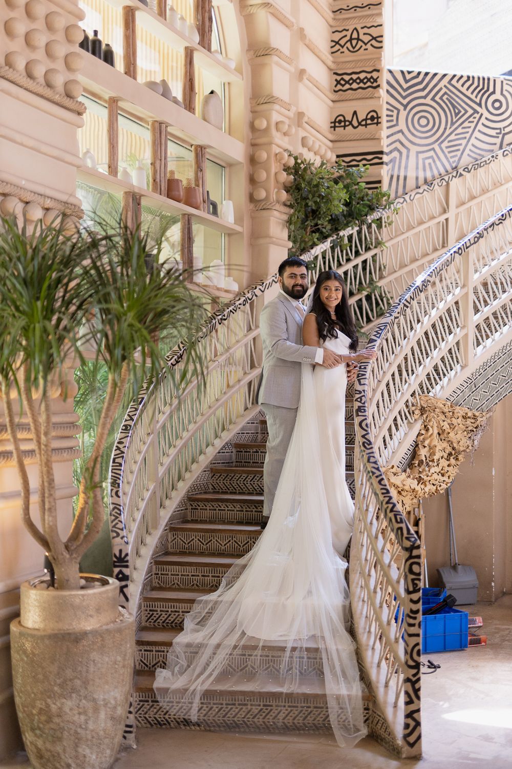 A couple poses on an ornate staircase with intricate metalwork railings and a flowing white wedding dress train.