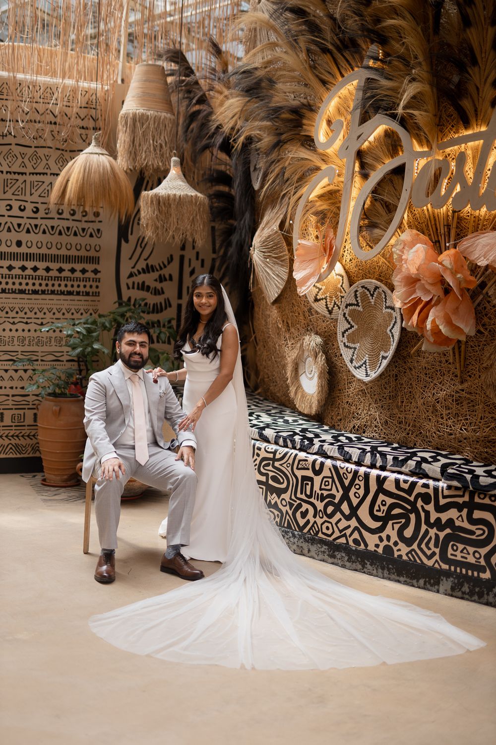 A couple in wedding attire poses against a decorative wall with ornate patterns and hanging lights in an elegant venue.