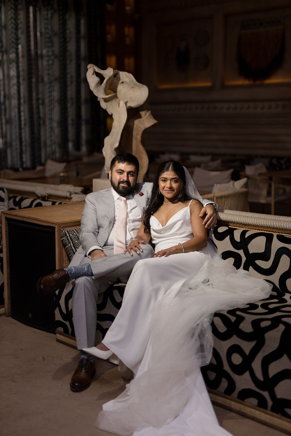 A couple in wedding attire sits together on an elegant bench in a beautifully decorated indoor setting.