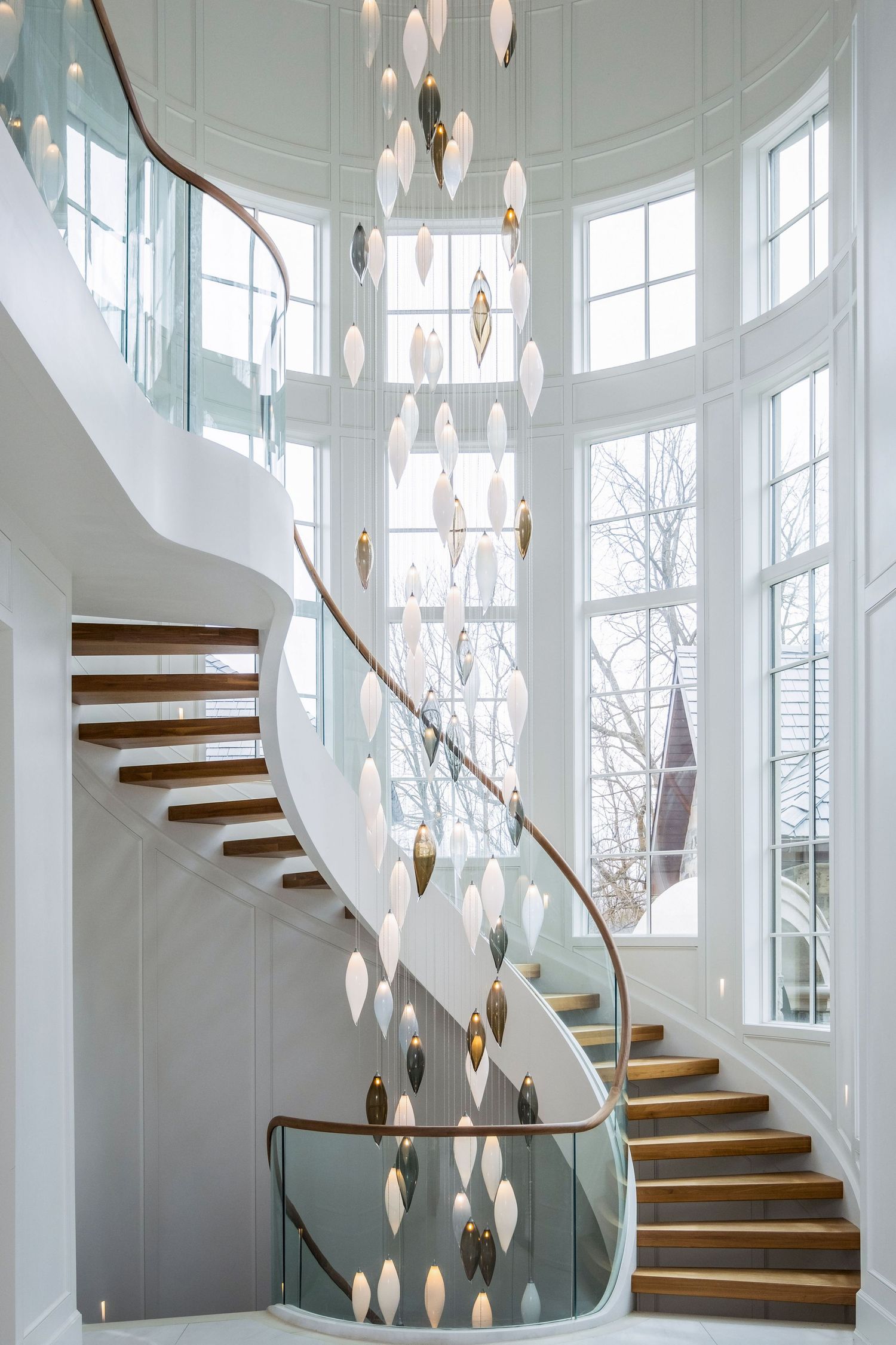 Elegant curved staircase with glass balustrade, wood steps, and cascading pendant chandelier in a bright white foyer.