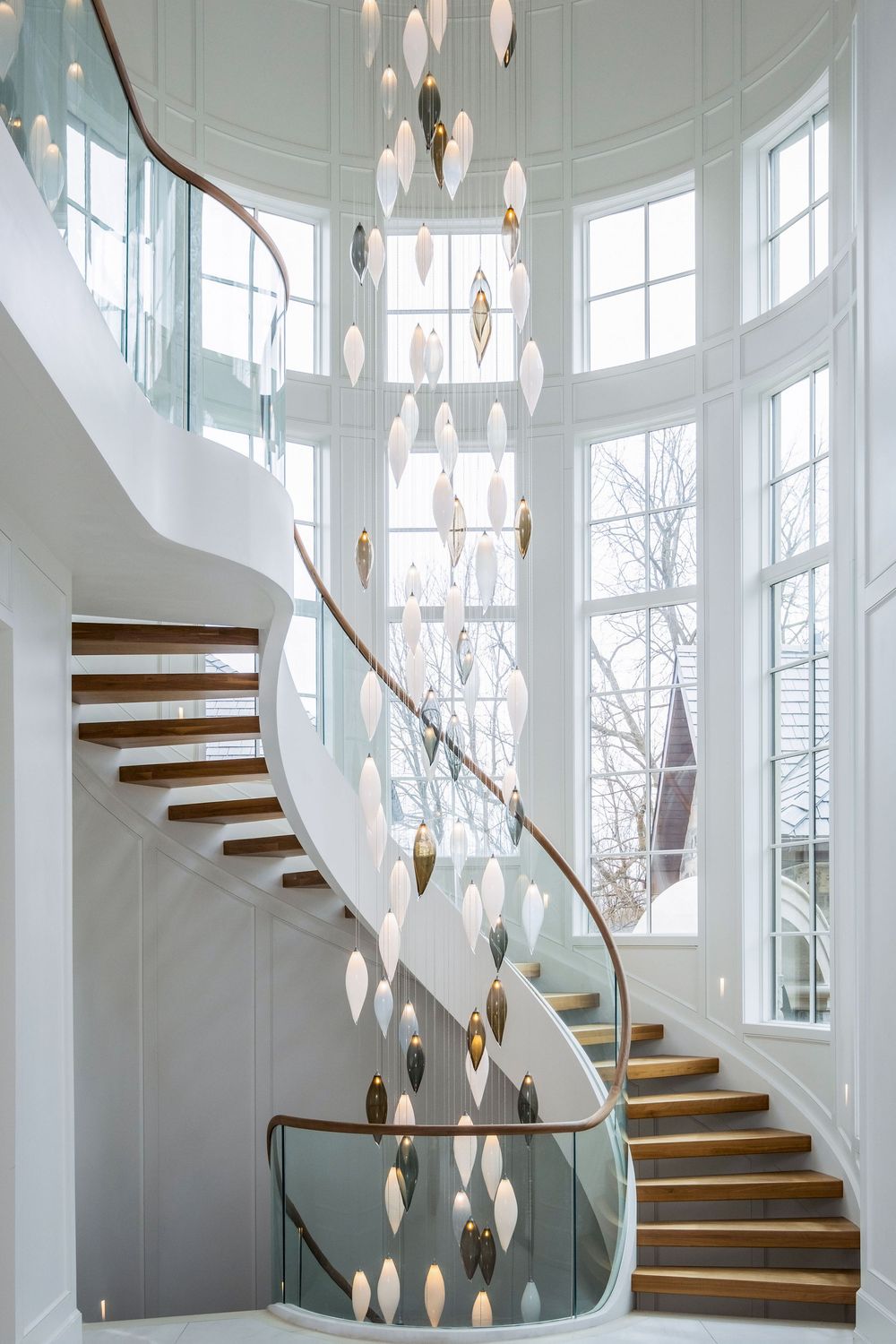 Elegant curved staircase with glass balustrade, wood steps, and cascading pendant chandelier in a bright white foyer.