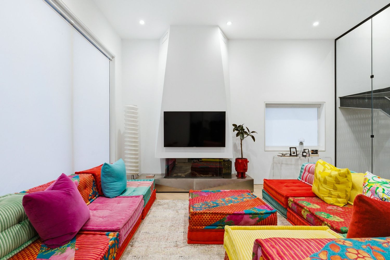 Modern living room with vibrant red and pink floor cushions, colorful throw pillows, and wall-mounted TV in a white space.