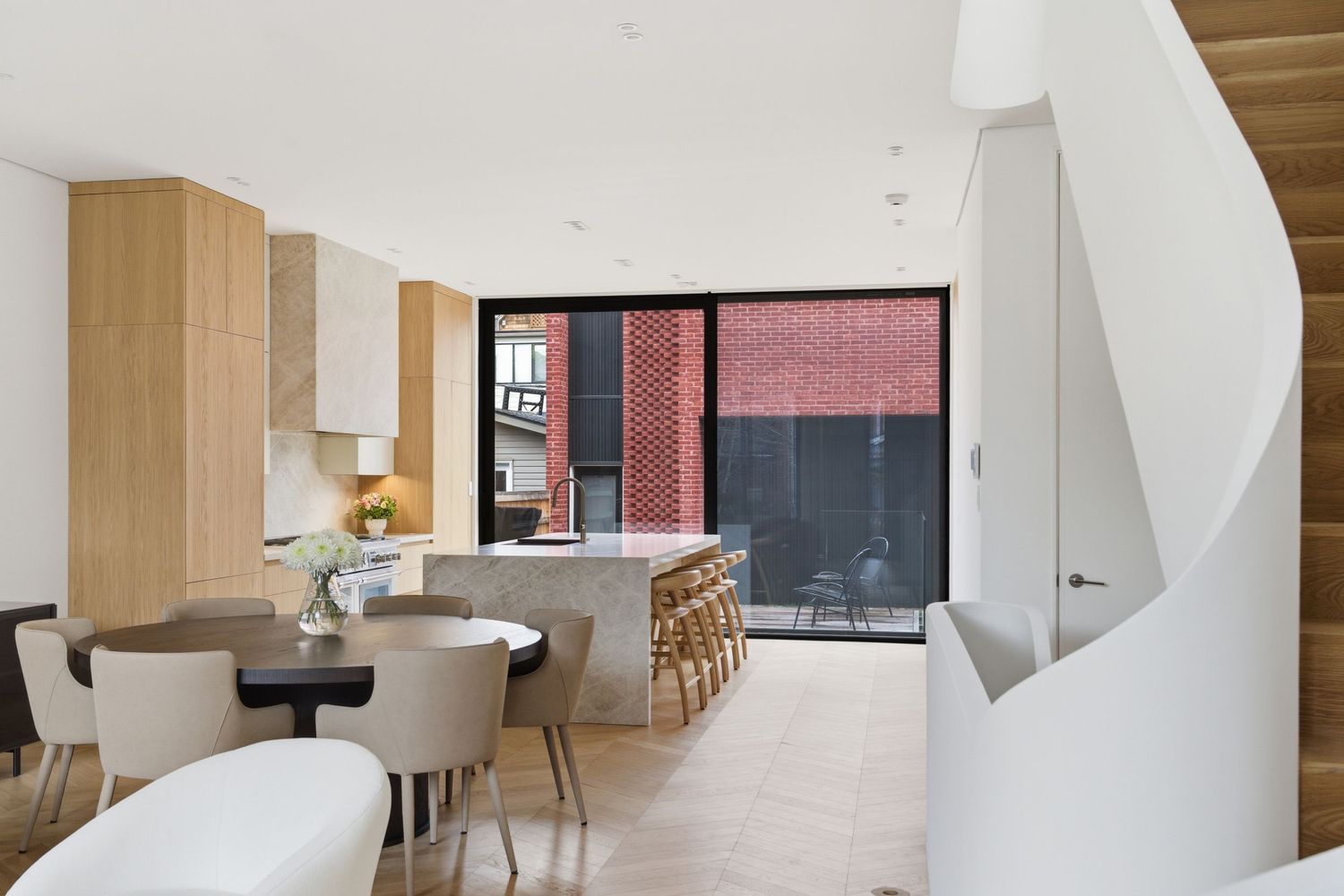 Modern open-plan kitchen and dining area with marble island, light wood cabinets, and floor-to-ceiling windows overlooking brick exterior.