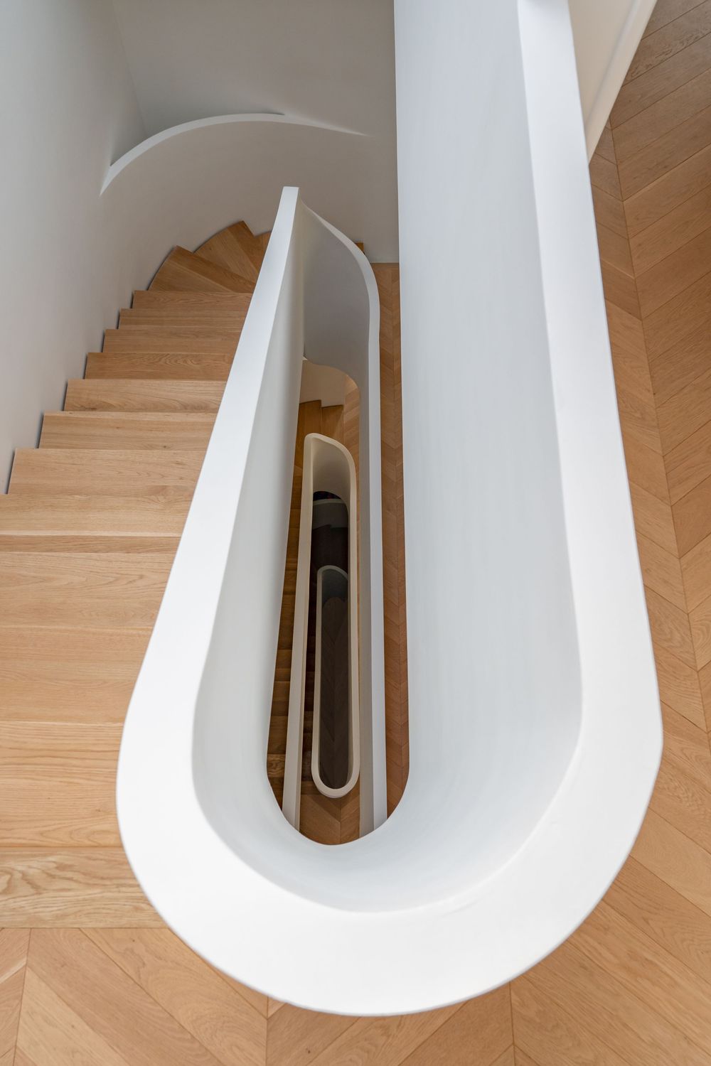 Aerial view of a modern spiral staircase with white curved railings and warm oak wood steps in a contemporary interior.