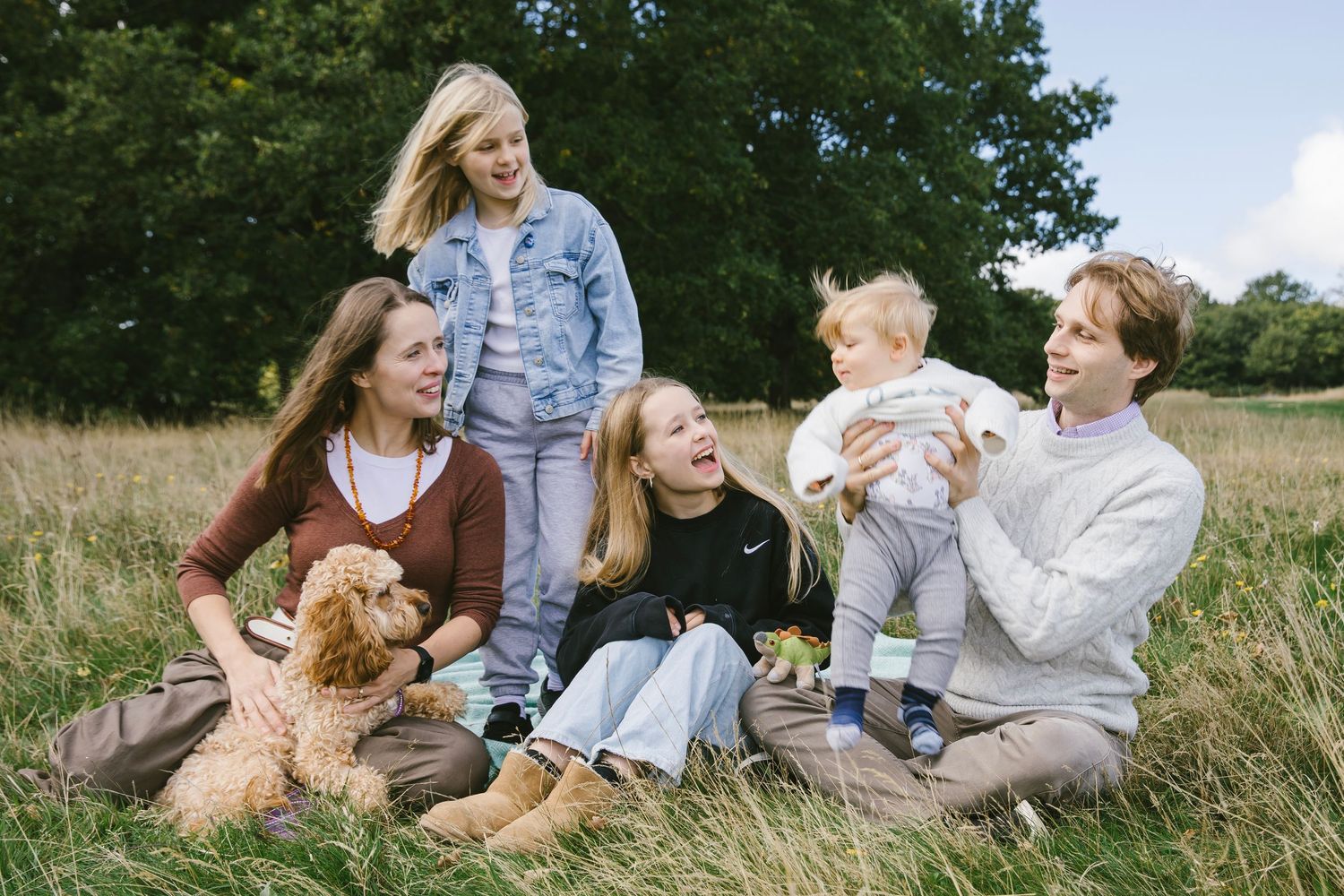Happy family with two girls, a baby, and a golden doodle dog sitting in a sunny meadow surrounded by trees.