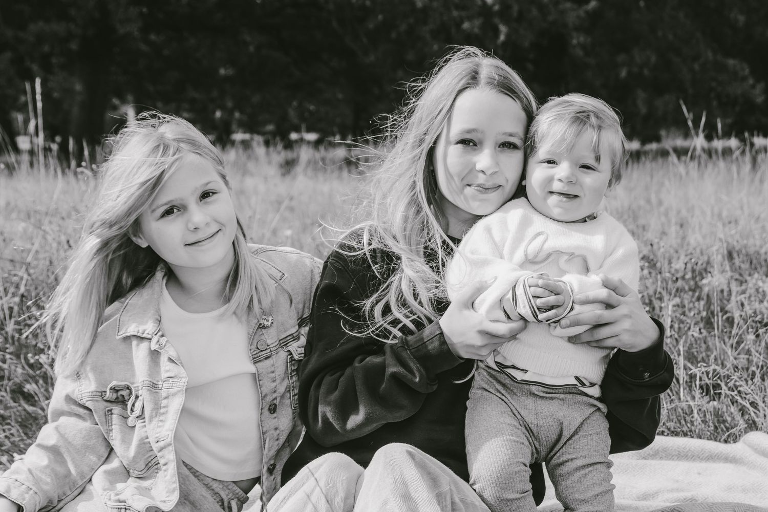 Black and white photo of a woman sitting outdoors with two young children, smiling in a grassy field.
