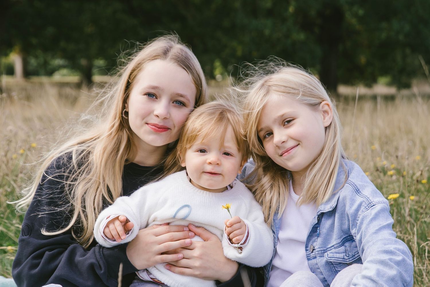 Three sisters sitting together in a sunny meadow, the two older girls hugging their baby sibling.