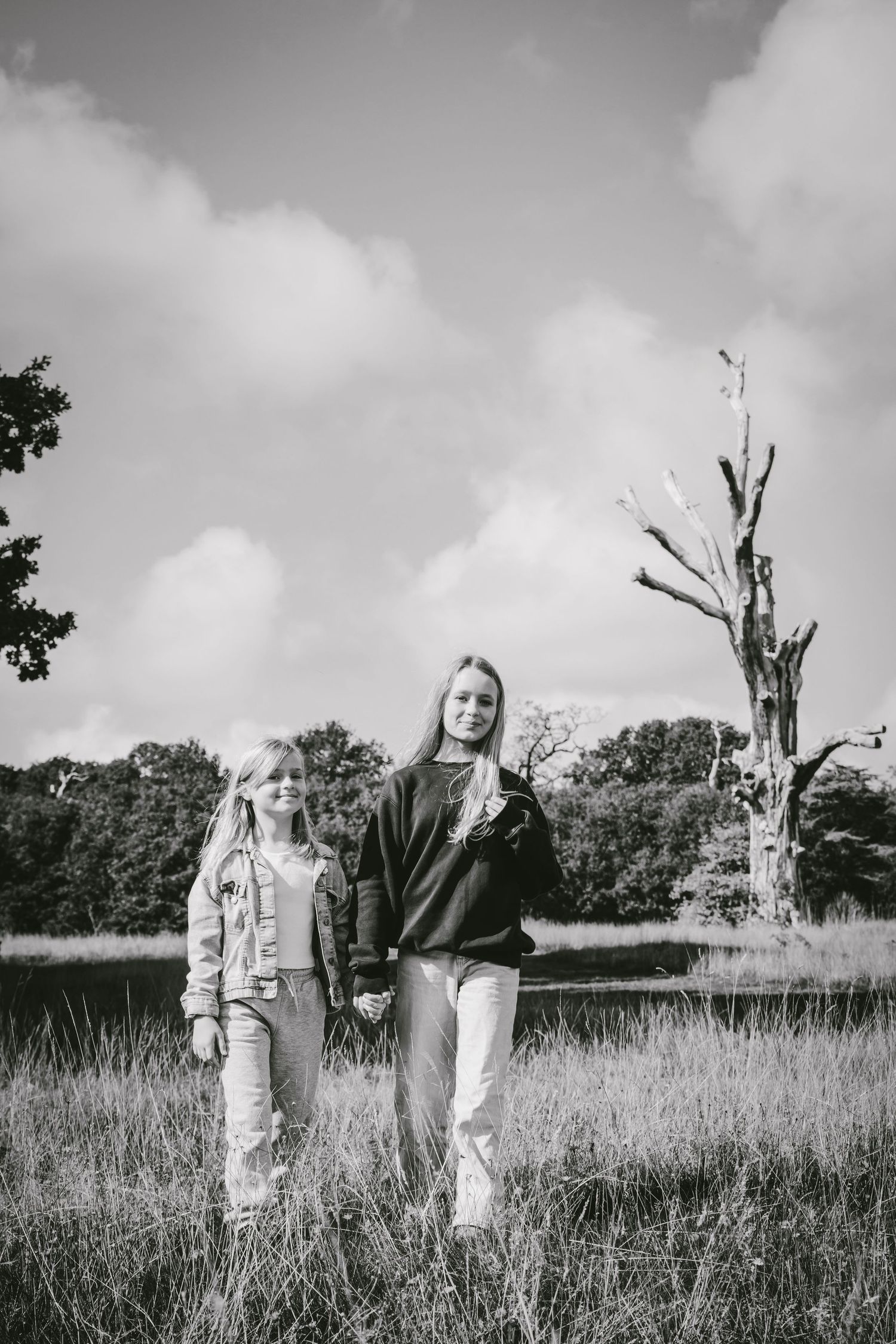 Two girls walking hand in hand through a grassy field with trees and a pond in a black and white photo.