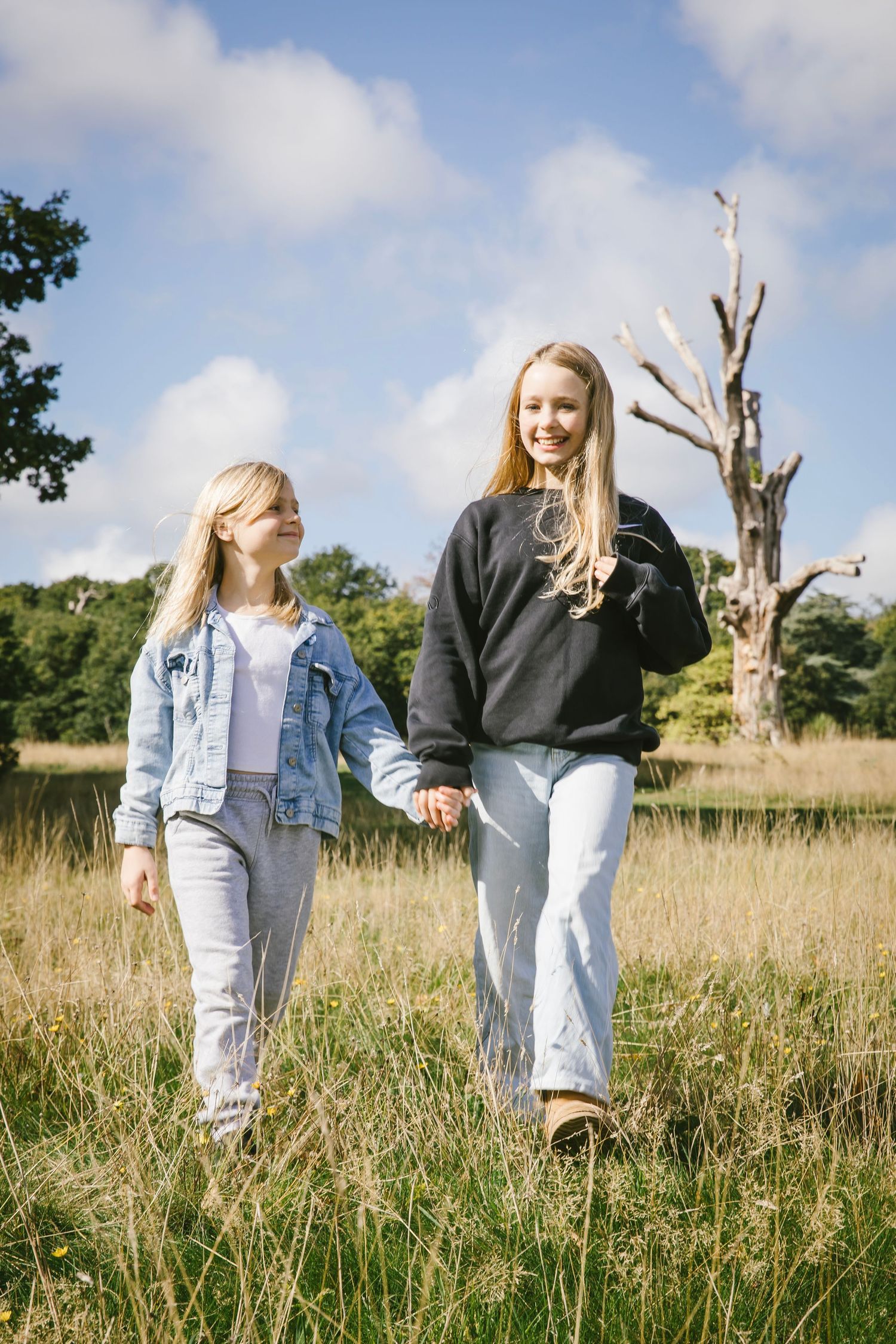 Two blonde girls walk hand-in-hand through a sunny meadow, wearing casual denim outfits with trees and blue sky behind.