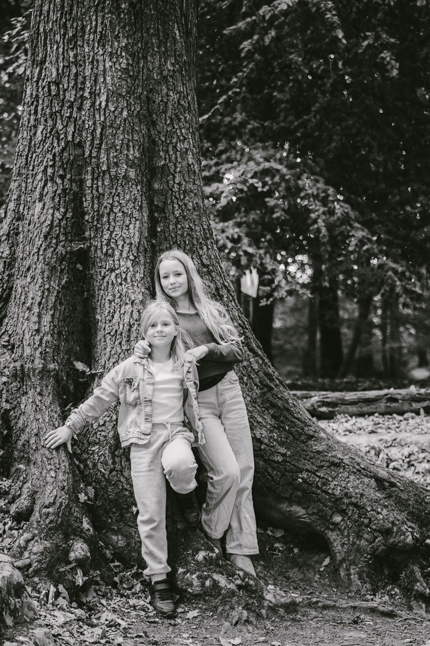 Black and white photo of two girls posing against a large tree trunk in a forest setting.