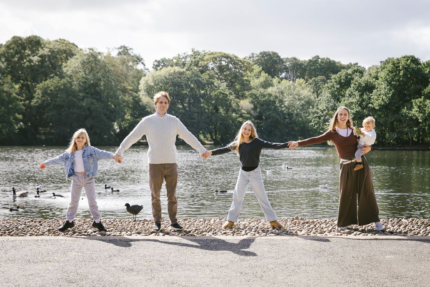 Family of five holding hands and stretching apart playfully by a scenic lake outdoors.