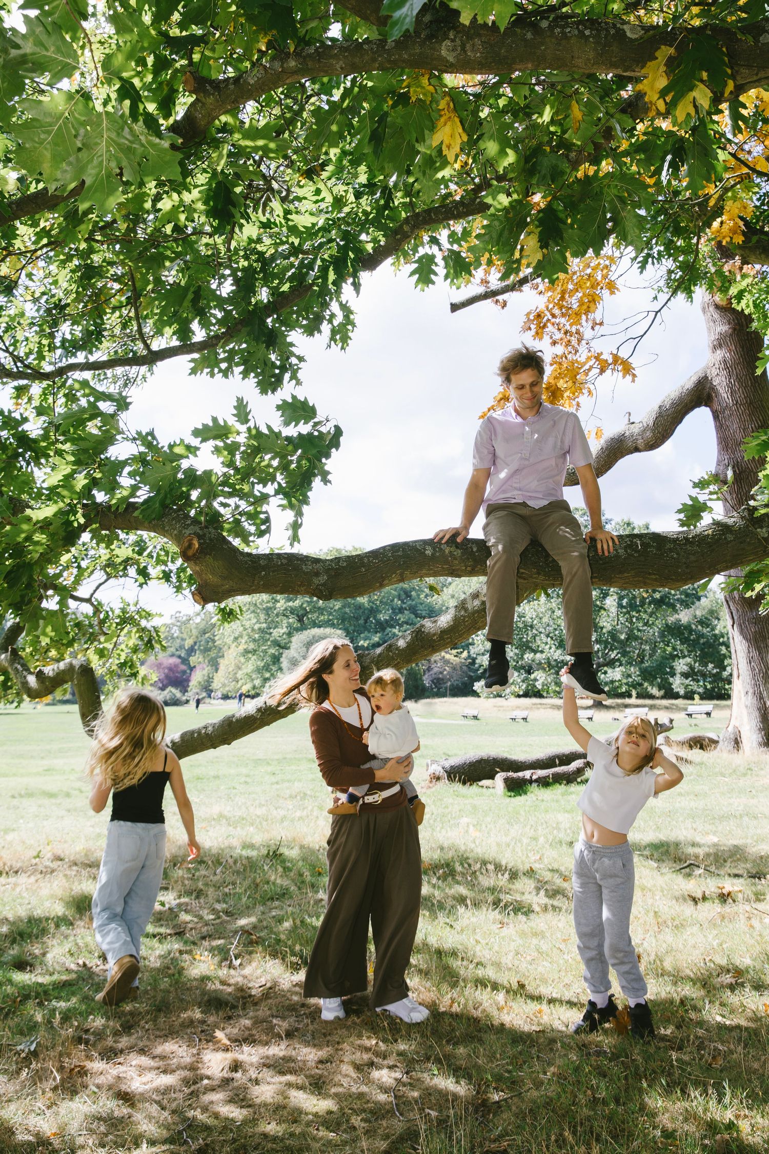 Family enjoying outdoor fun with man sitting in tree branches while woman holds baby and children play.