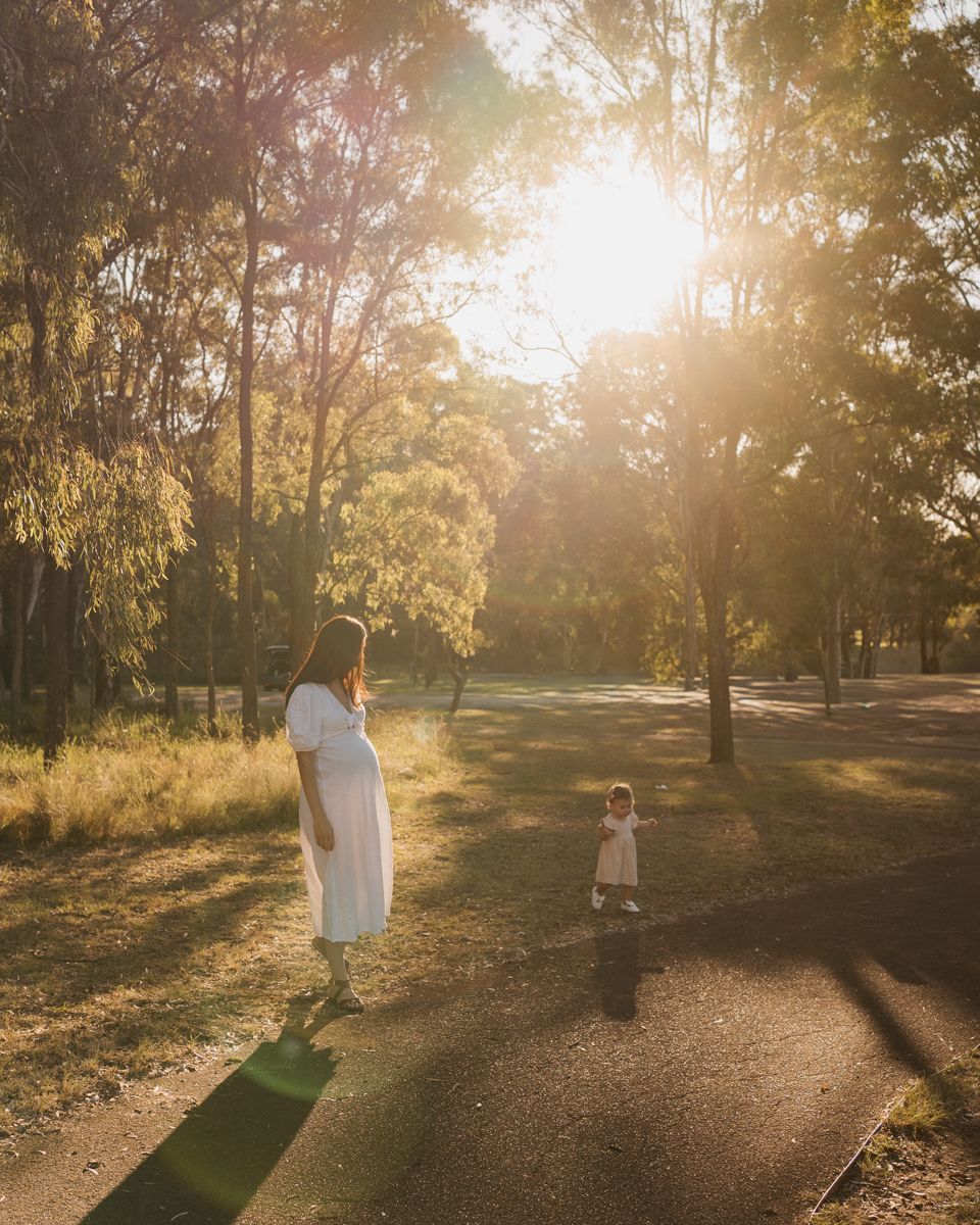 Pregnant woman in white dress stands with toddler on sunlit park path surrounded by tall trees at golden hour rouse hill family photoshoot