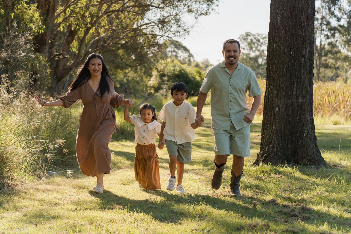 Happy family of four running and holding hands in a sunny green park with trees in the background during family photoshoot in Rouse hill