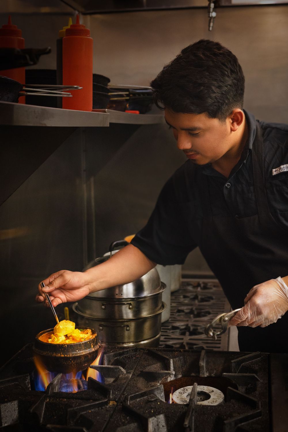 Chef cooking vibrant dish in a clay pot over open flame on a commercial gas stove in a restaurant kitchen.