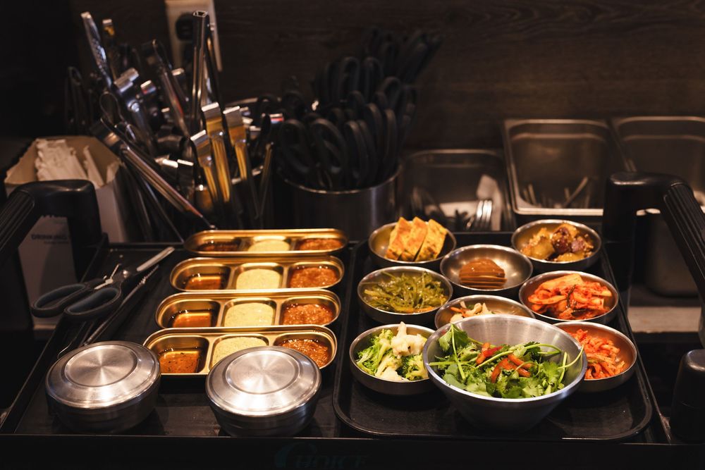 Korean banchan spread with colorful side dishes in metal bowls and trays on a dark restaurant counter.